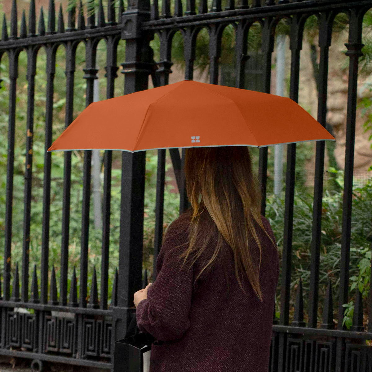 Woman walking down the street while holding an opened Weatherman Travel Umbrella in rusty orange.