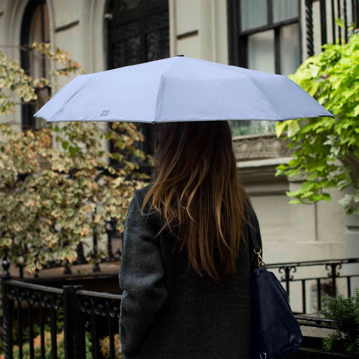 Woman walking down the street while holding an opened Weatherman Travel Umbrella in whispy blue.
